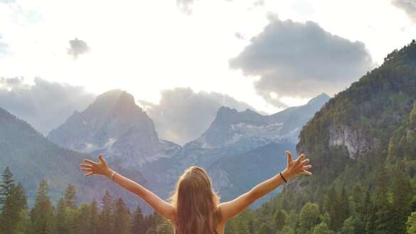 Junge Frau steht an einem See, im Hintergrund Berge. Sie hat die Arme ausgebreitet